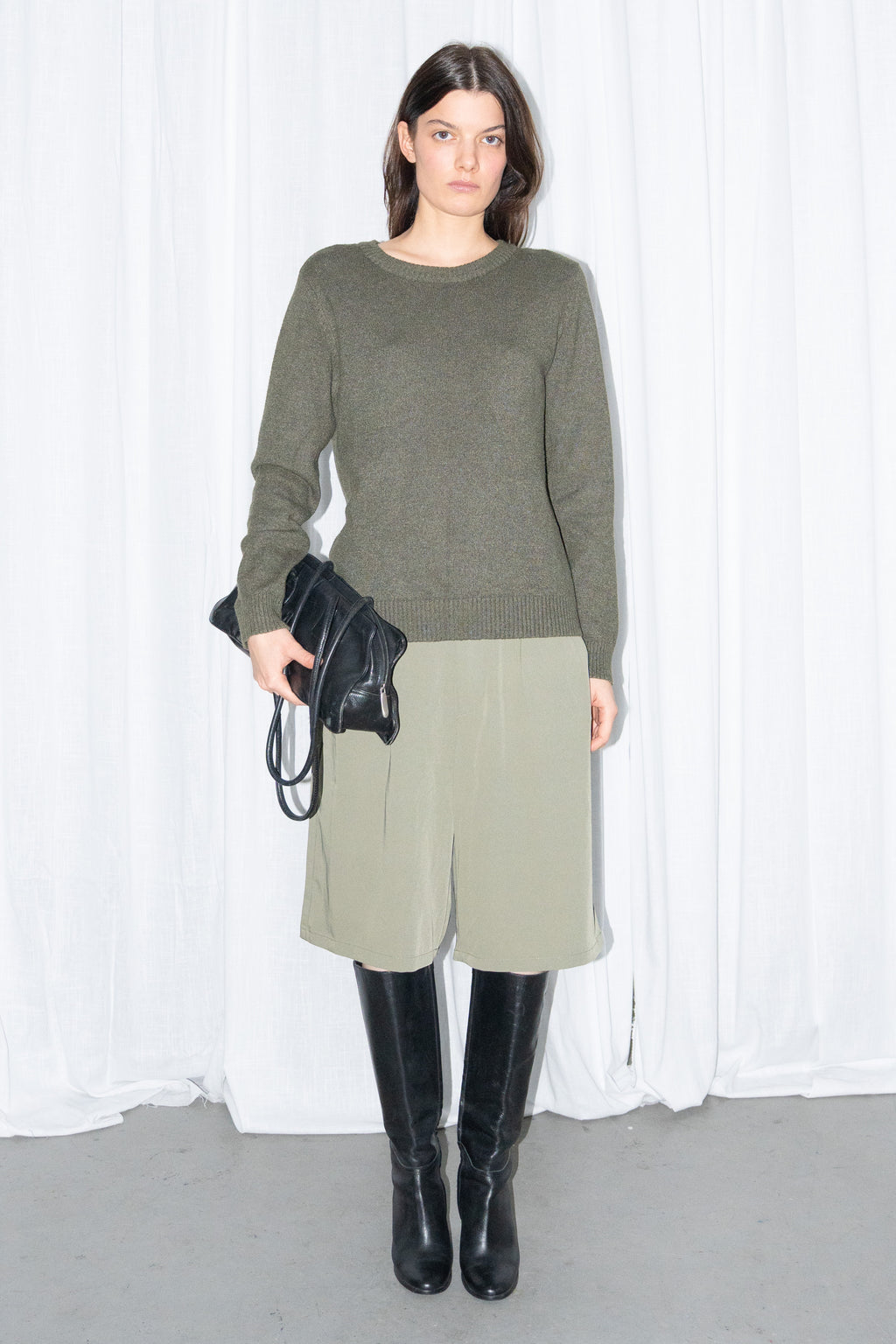 Model wearing a dark olive green crew-neck knit sweater with khaki green tailored Bermuda shorts and black knee boots, holding a black bag