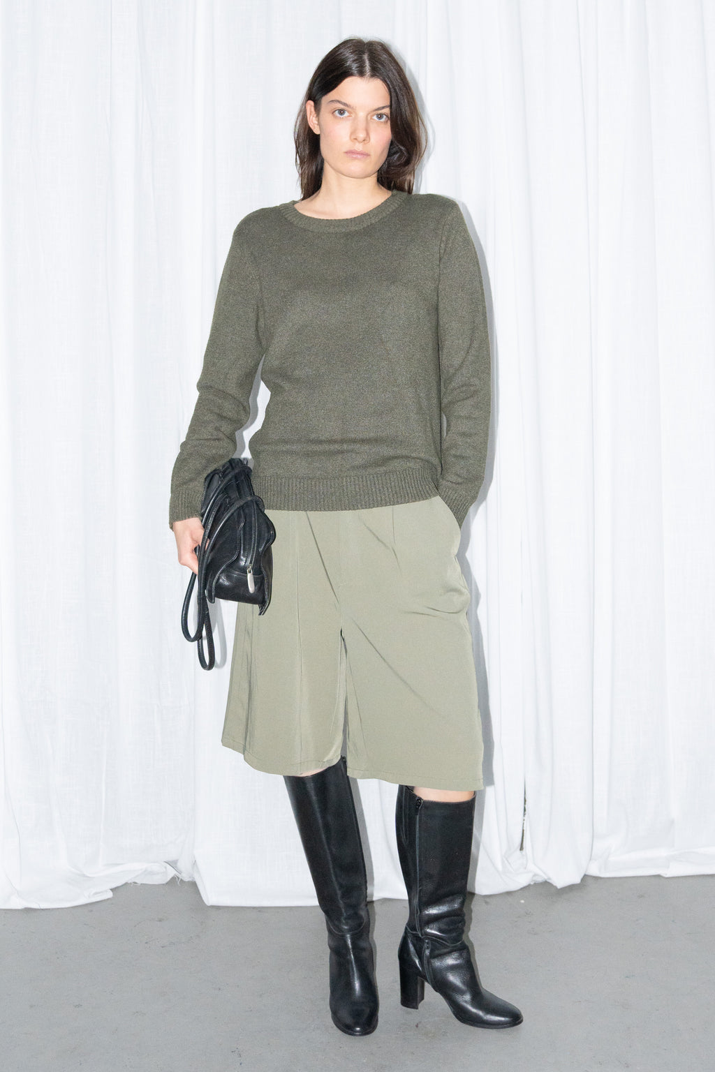 Model in a dark olive green crew-neck knit sweater and khaki green Bermuda shorts, holding a black bag