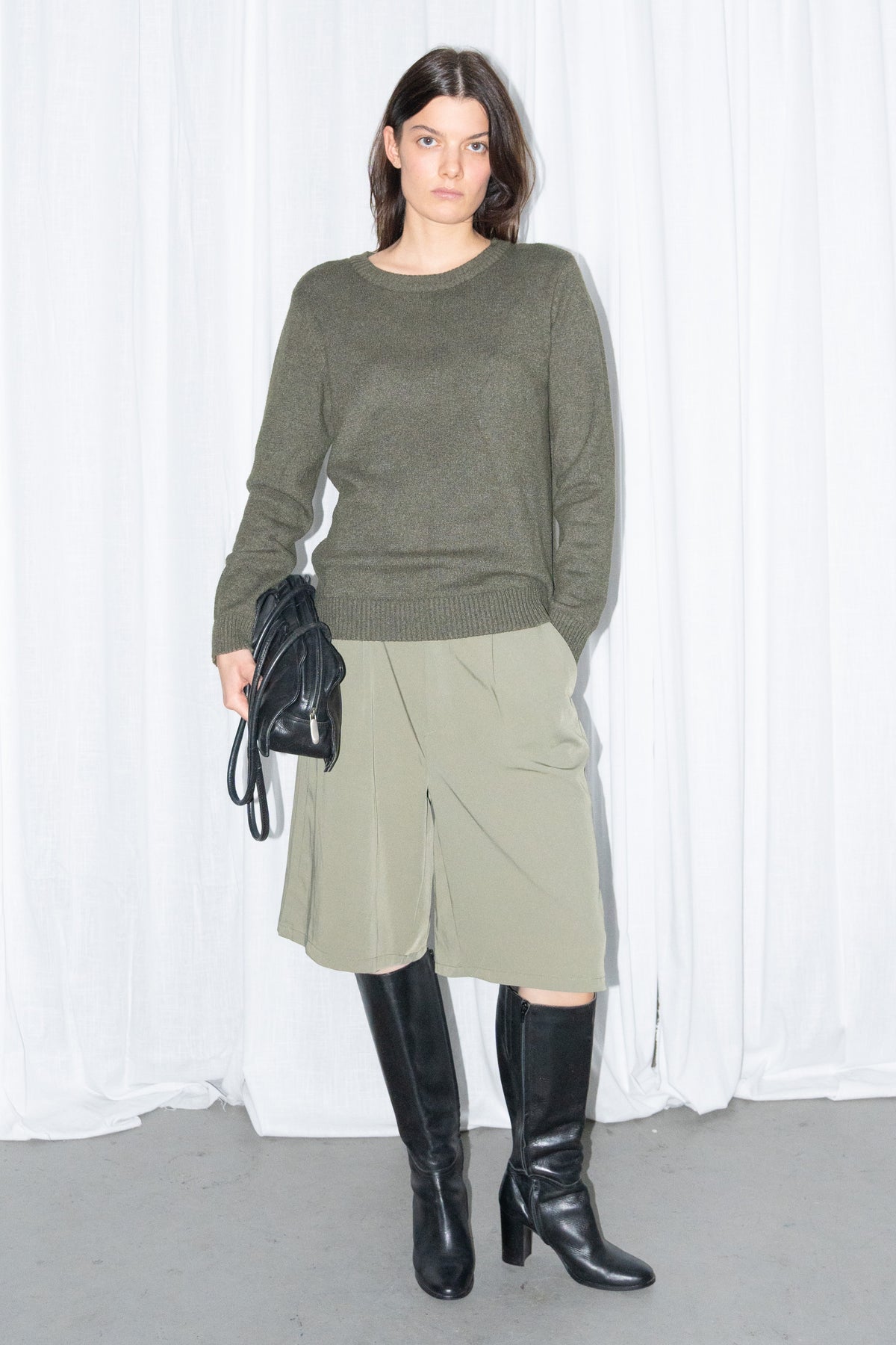 Model in a dark olive green crew-neck knit sweater and khaki green Bermuda shorts, holding a black bag