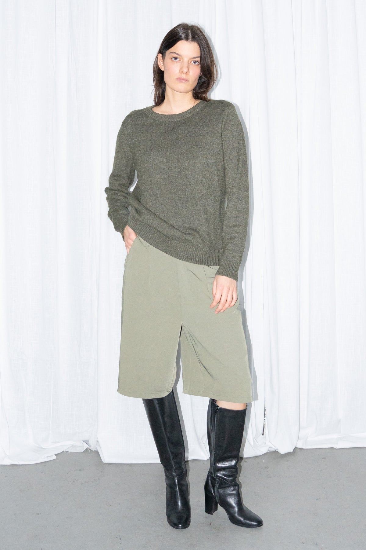 Model posing in a dark olive green crew-neck knit sweater and khaki green Bermuda shorts