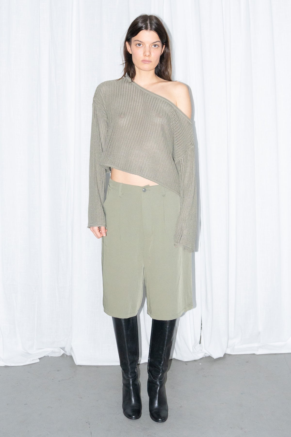Model wearing an olive off-shoulder chunky knit sweater with khaki green tailored Bermuda shorts and black knee boots