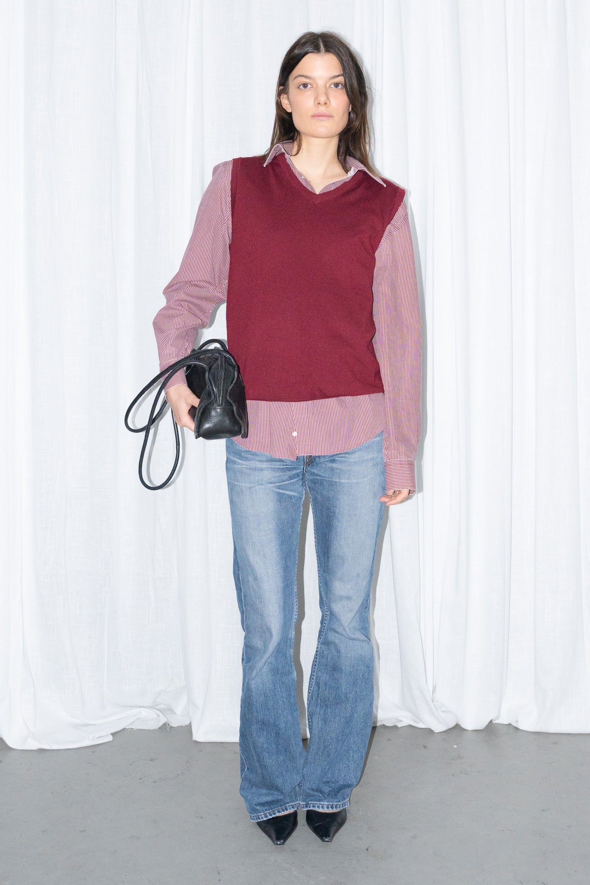 Model wearing a burgundy V-neck knit sweater vest over a burgundy striped button-up shirt with light blue jeans
