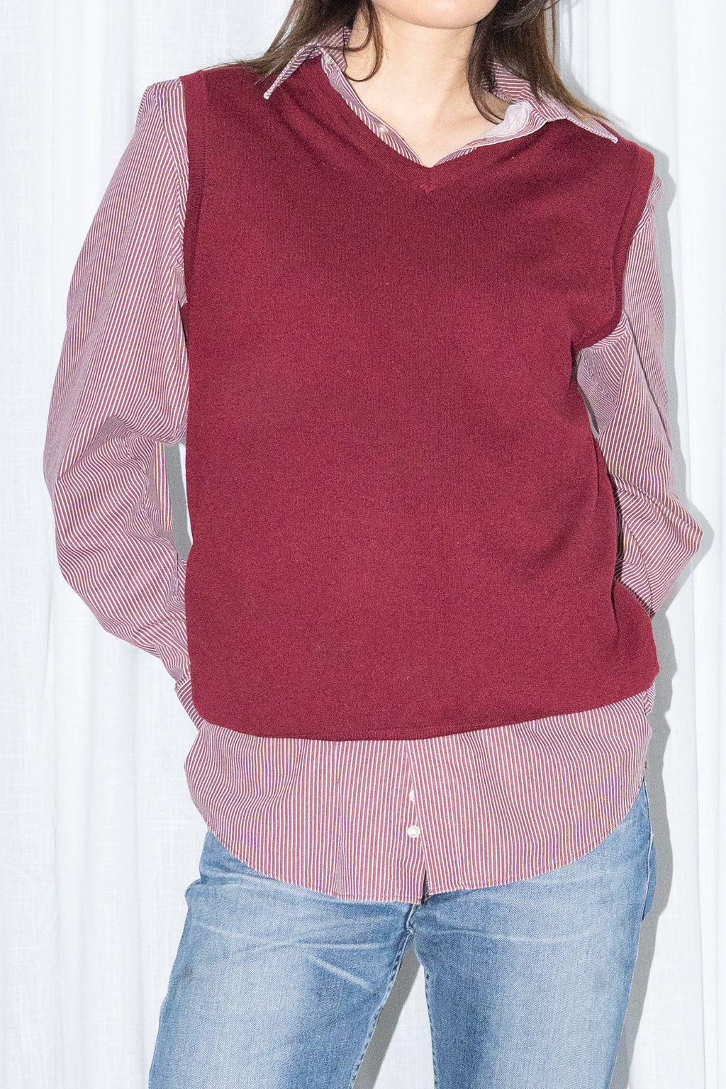 Close-up of a burgundy V-neck knit sweater vest over a burgundy striped button-up shirt with light blue jeans