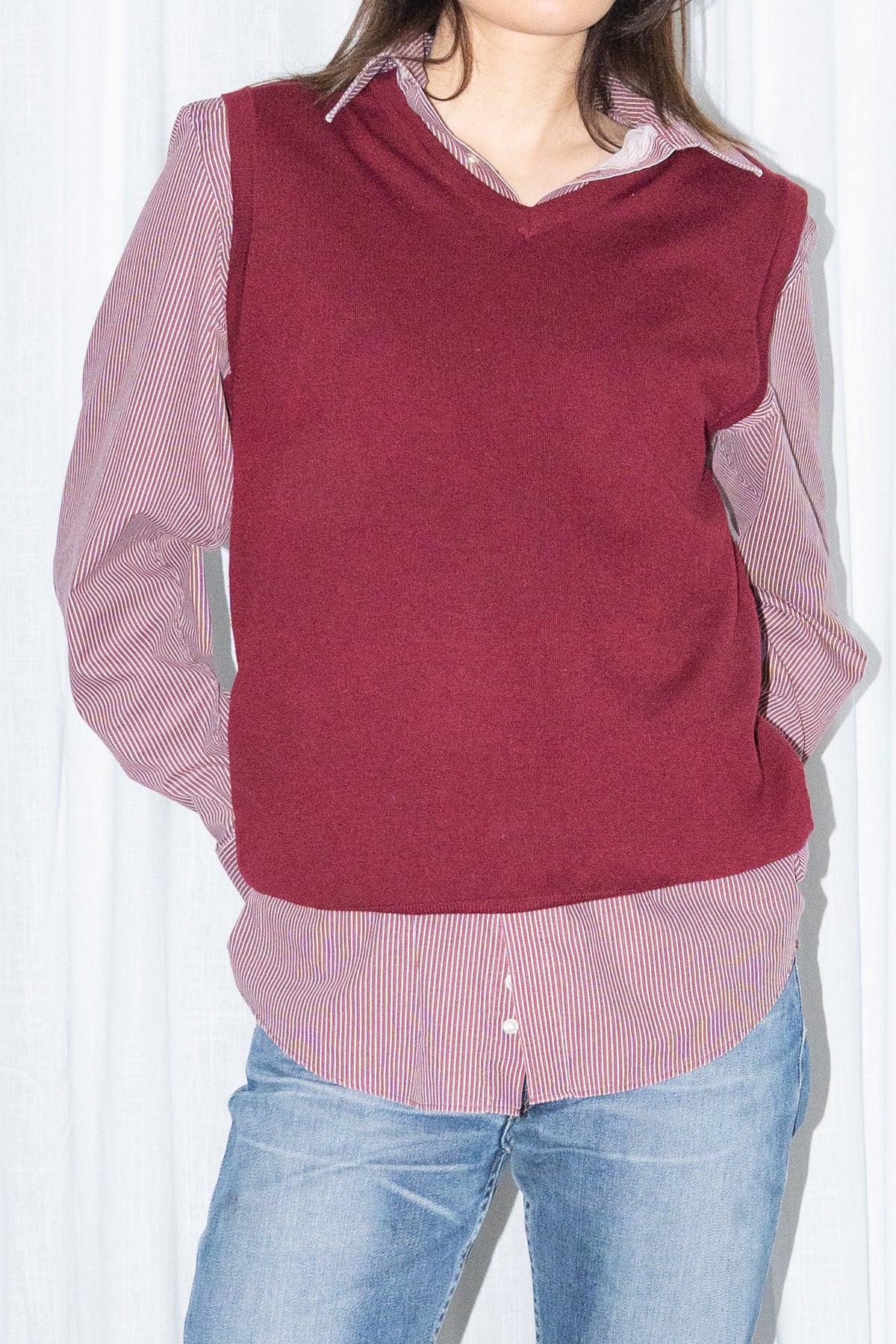 Close-up of a burgundy V-neck knit sweater vest over a burgundy striped button-up shirt with light blue jeans