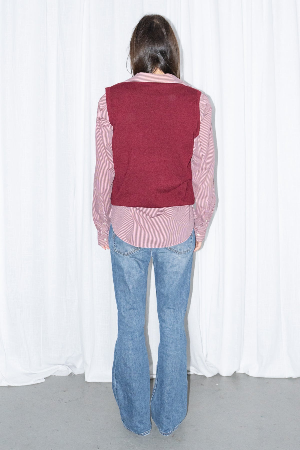 Back view of a burgundy V-neck knit sweater vest over a burgundy striped button-up shirt with light blue jeans