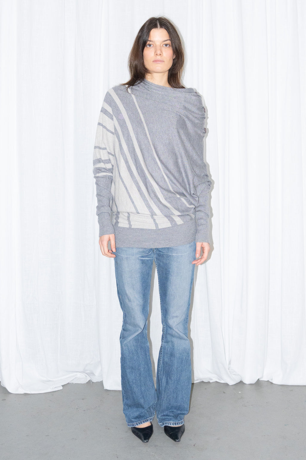 Model wearing a grey draped asymmetric knit top with light blue jeans