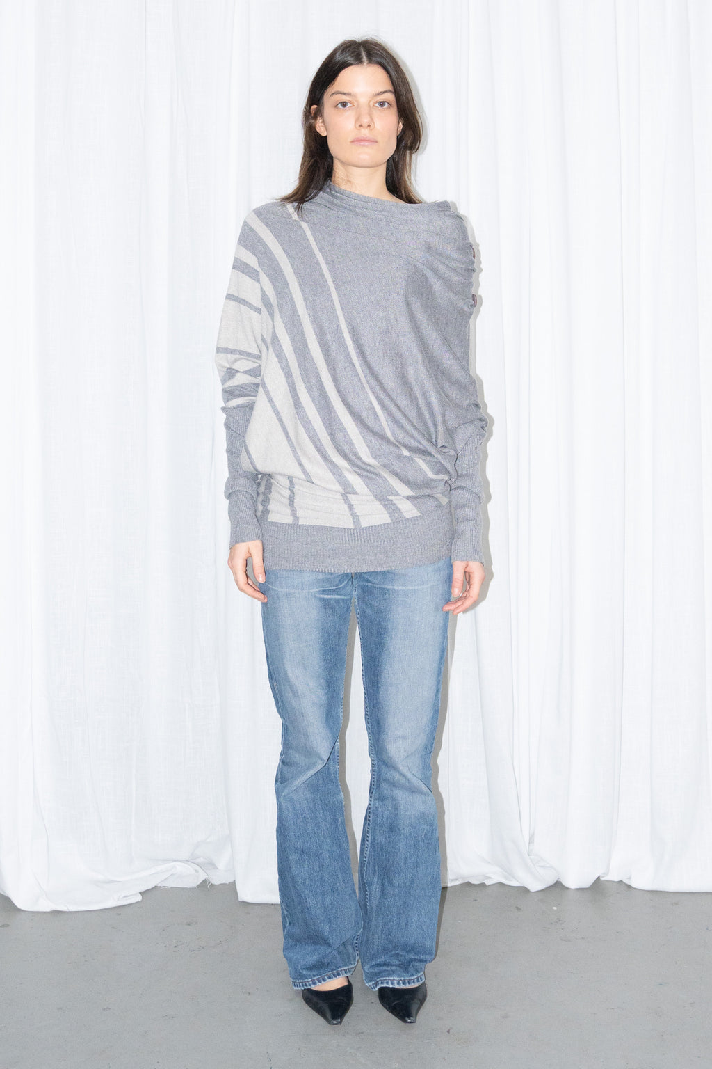 Model wearing a grey draped asymmetric knit top with light blue jeans