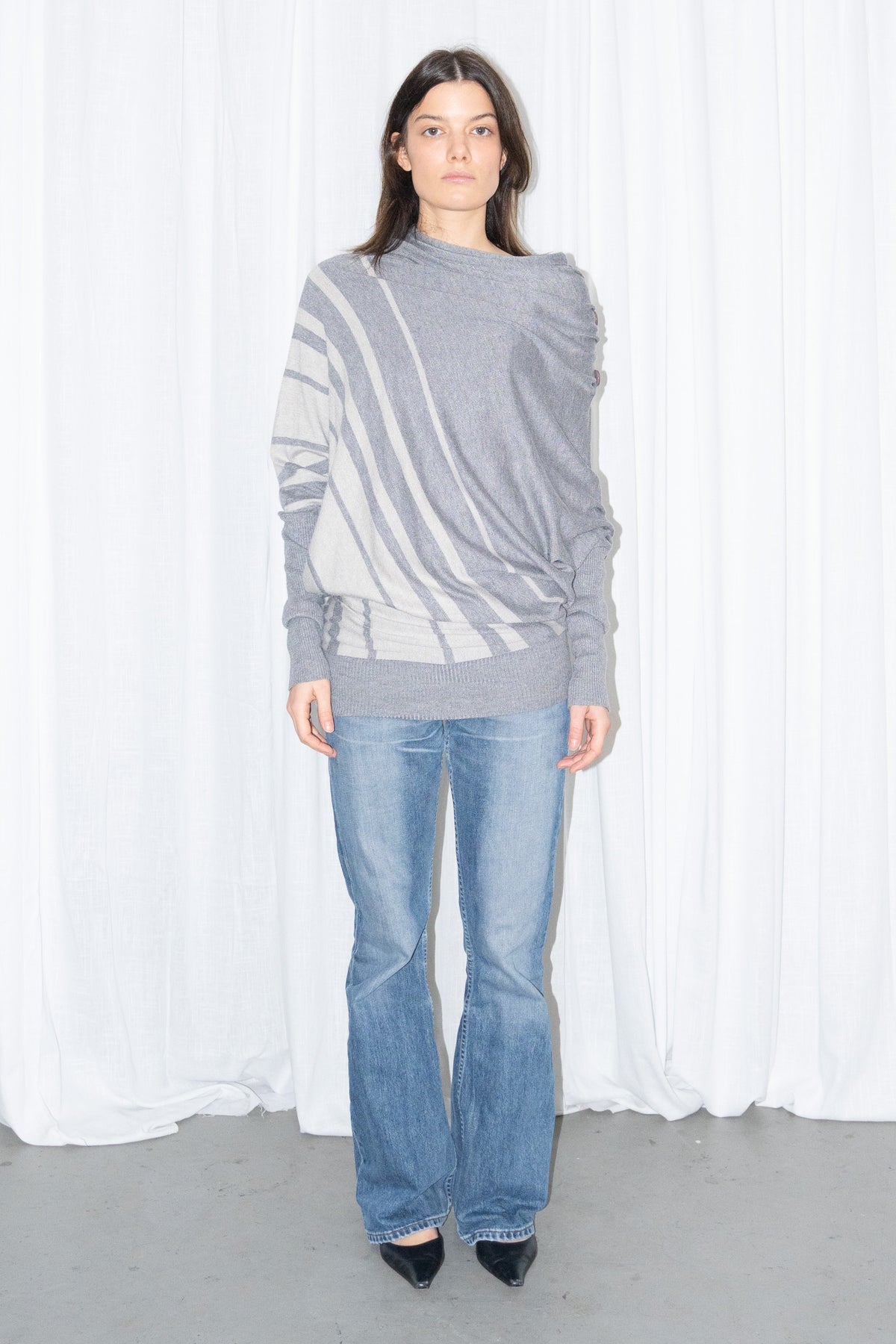 Model wearing a grey draped asymmetric knit top with light blue jeans