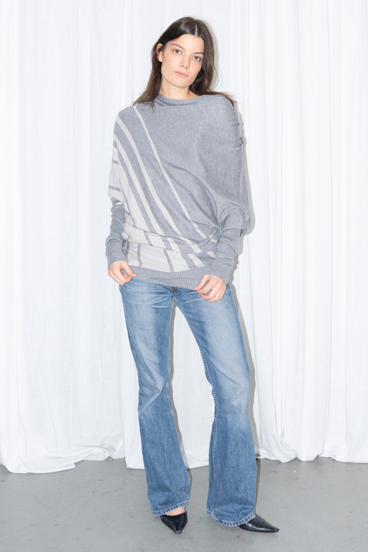 Model wearing a grey draped asymmetric knit top with light blue jeans