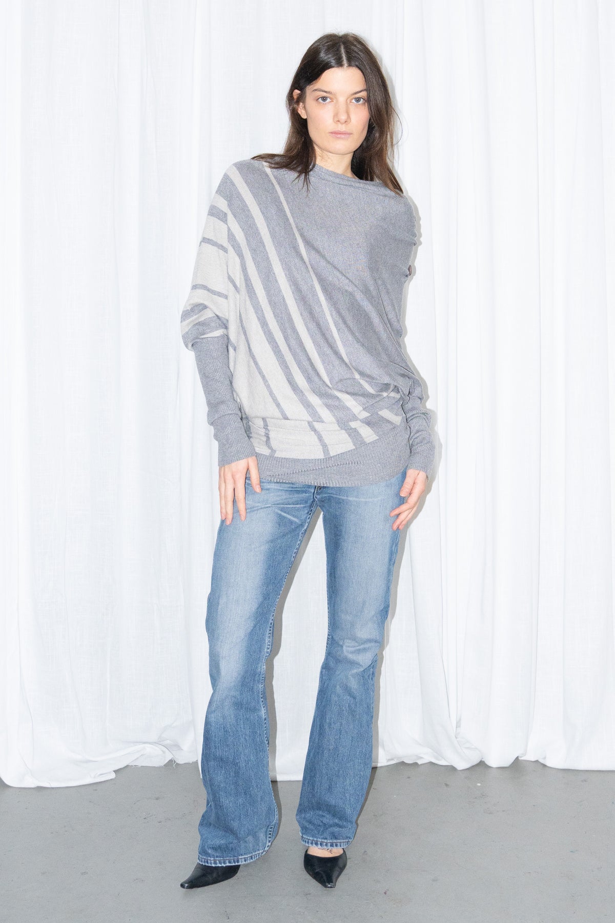 Model wearing a grey draped asymmetric knit top with light blue jeans