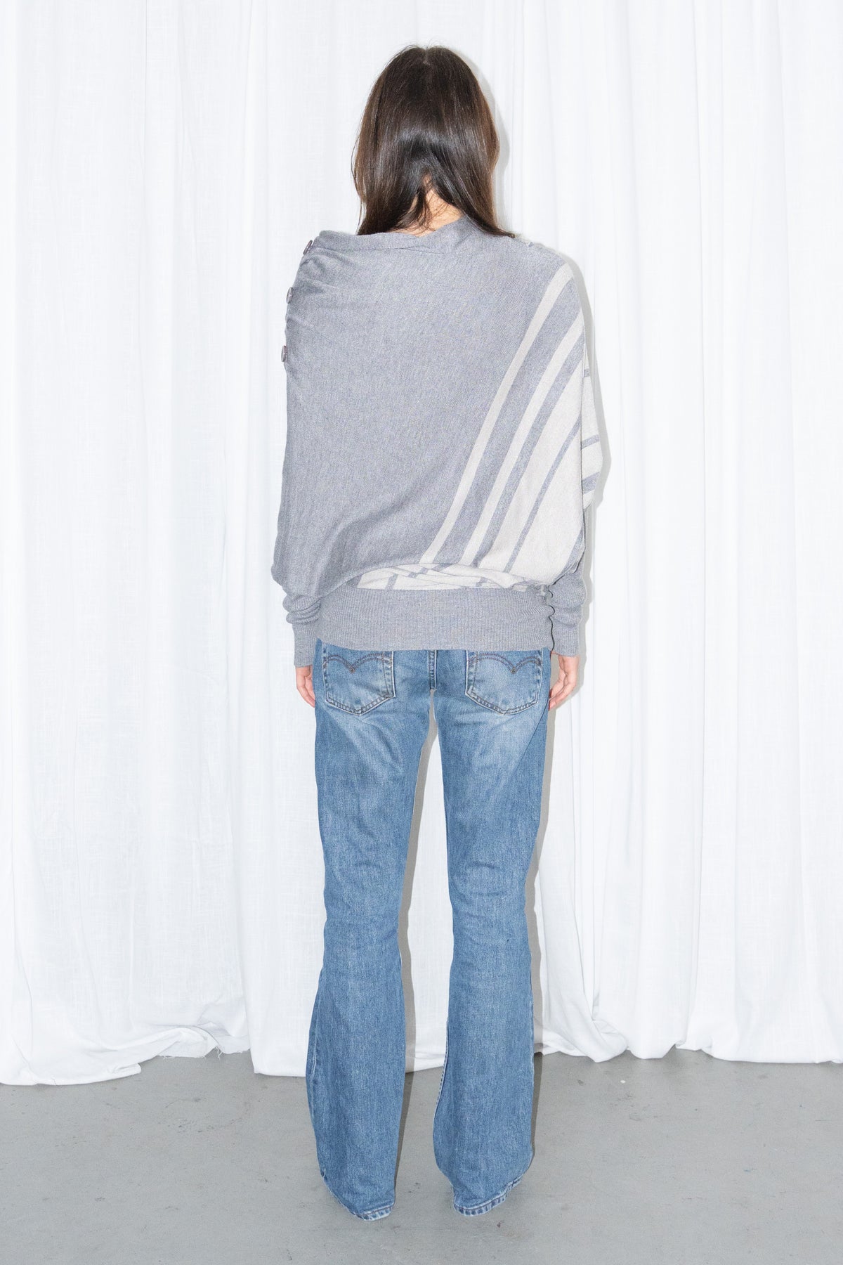 Back view of a grey draped asymmetric knit top with light blue jeans
