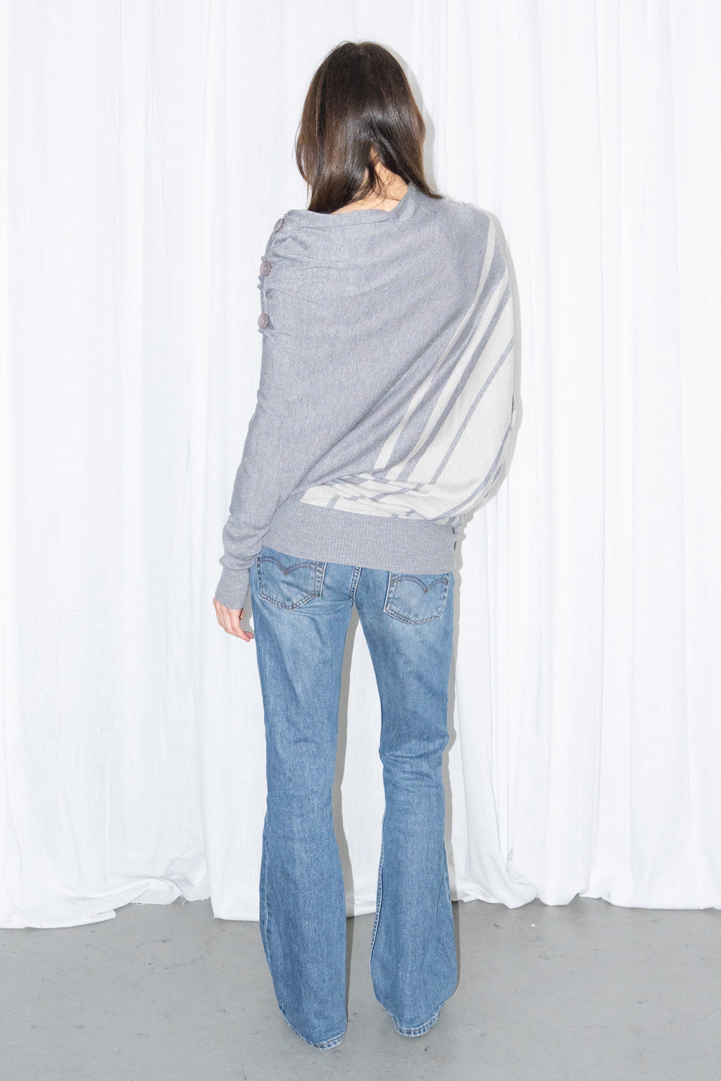 Back view of a grey draped asymmetric knit top with light blue jeans