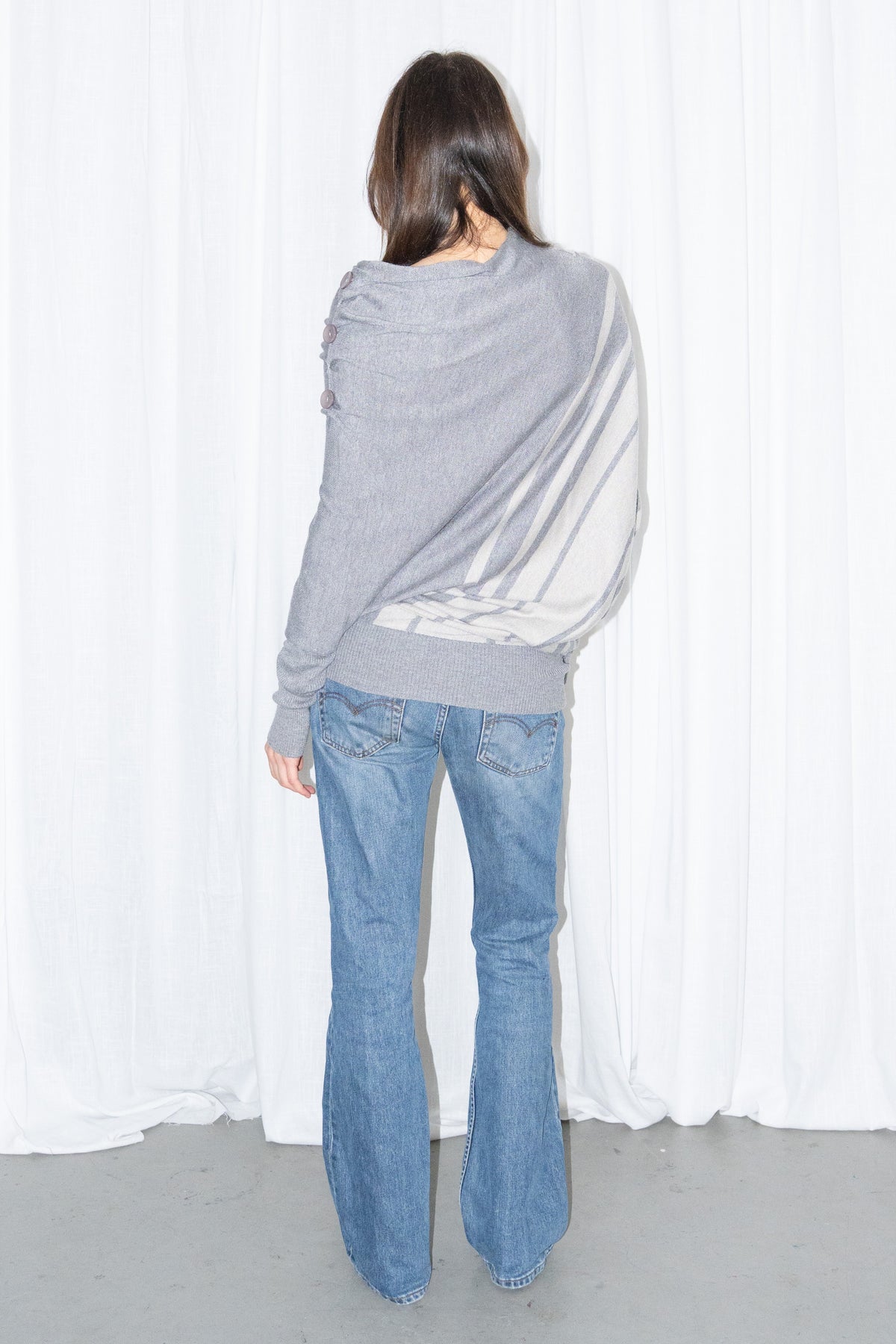 Back view of a grey draped asymmetric knit top with light blue jeans