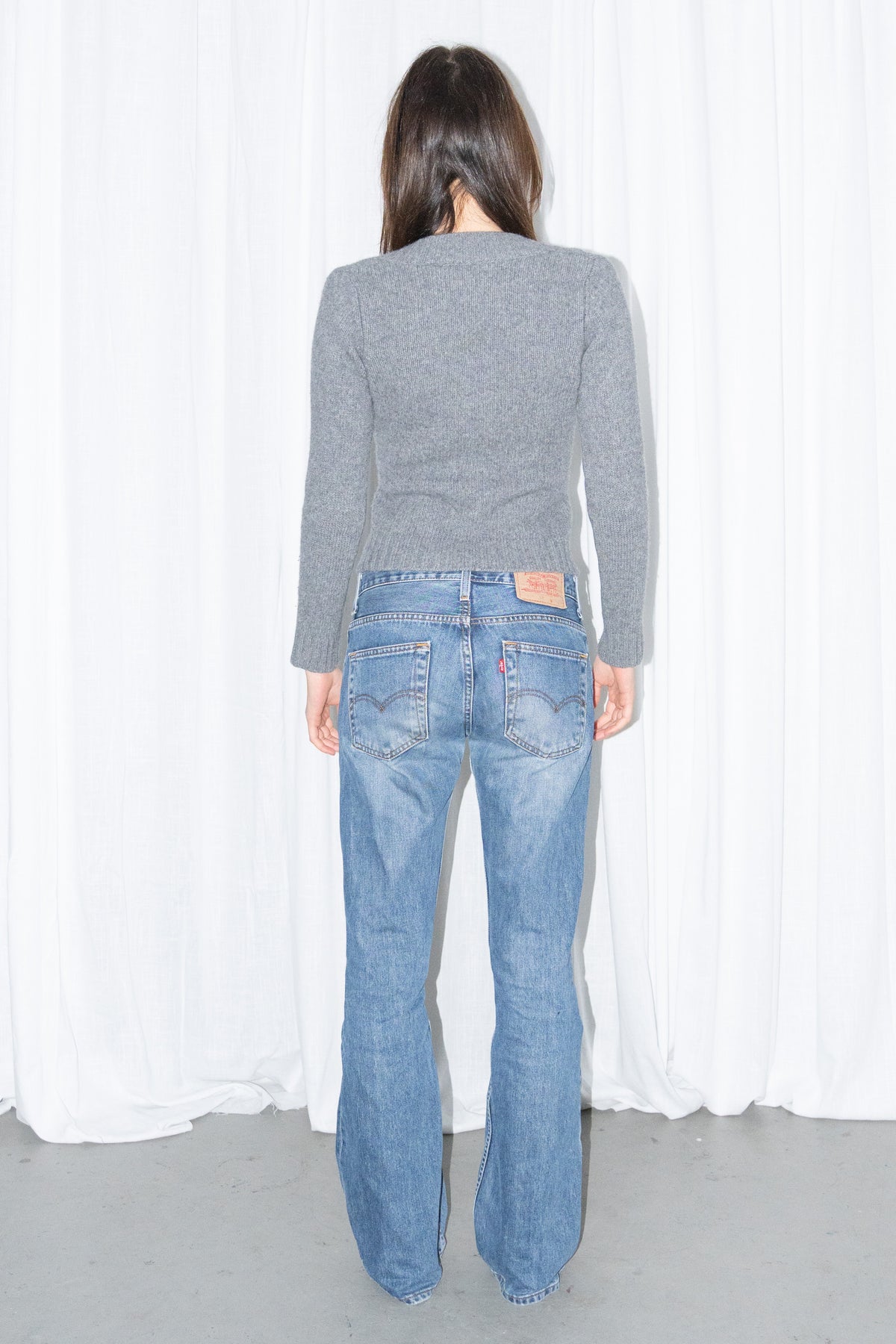 Back view of a grey deep V-neck cropped knit sweater with light blue jeans