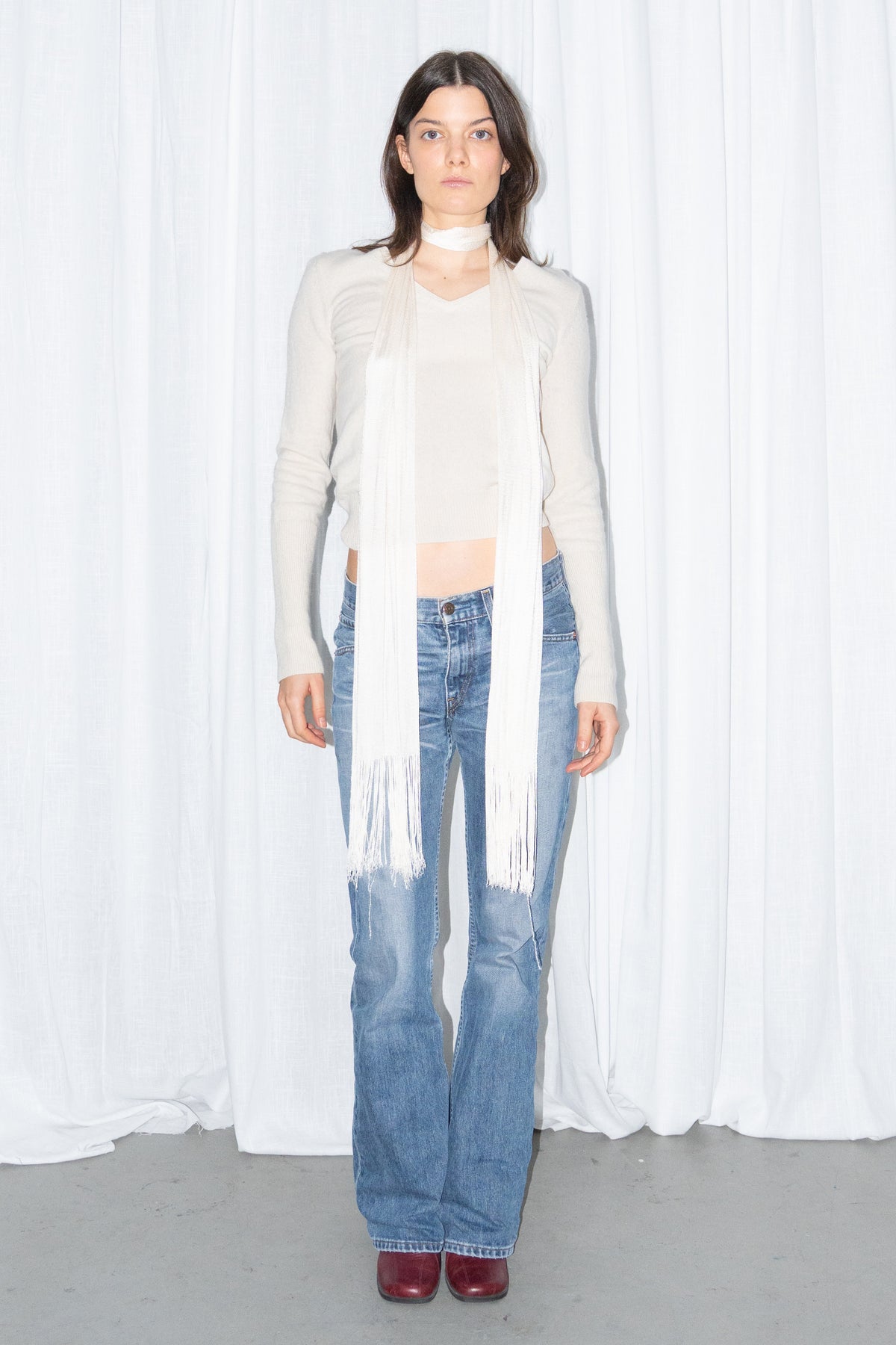 Model wearing a cream V-neck knit sweater and a long fringed scarf with light blue low-rise straight jeans