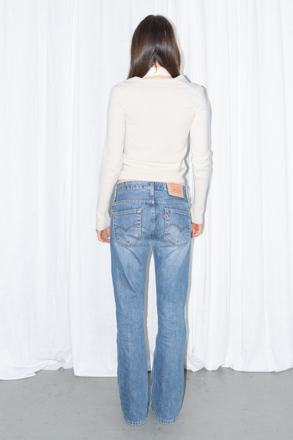 Back view of a cream V-neck knit sweater and a long fringed scarf with light blue low-rise straight jeans