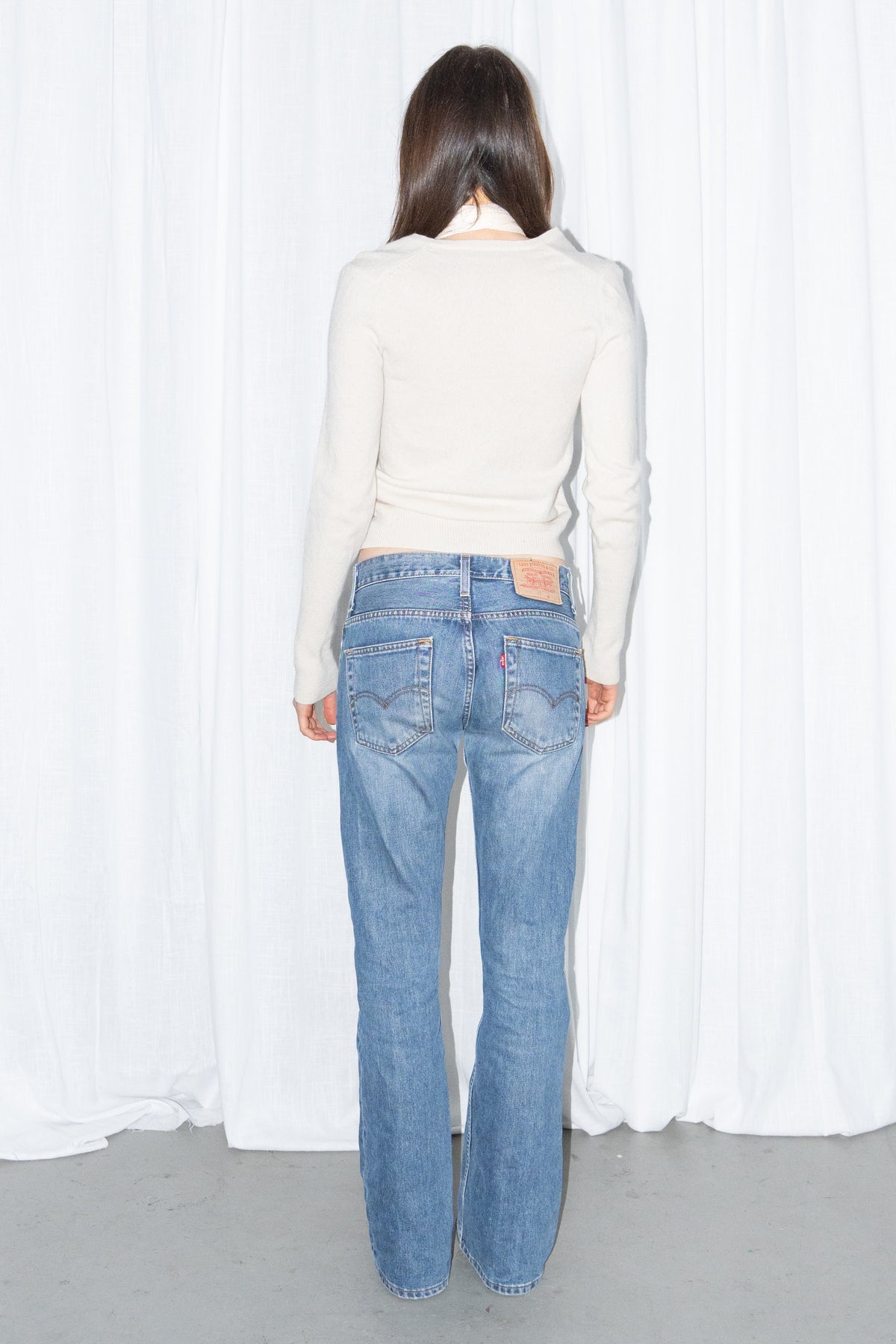 Back view of a cream V-neck knit sweater and a long fringed scarf with light blue low-rise straight jeans