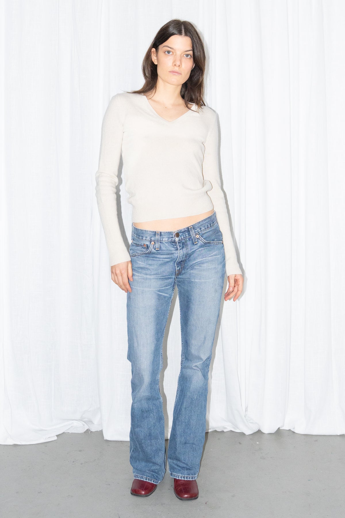 Model wearing a cream V-neck knit sweater and a long fringed scarf with light blue low-rise straight jeans