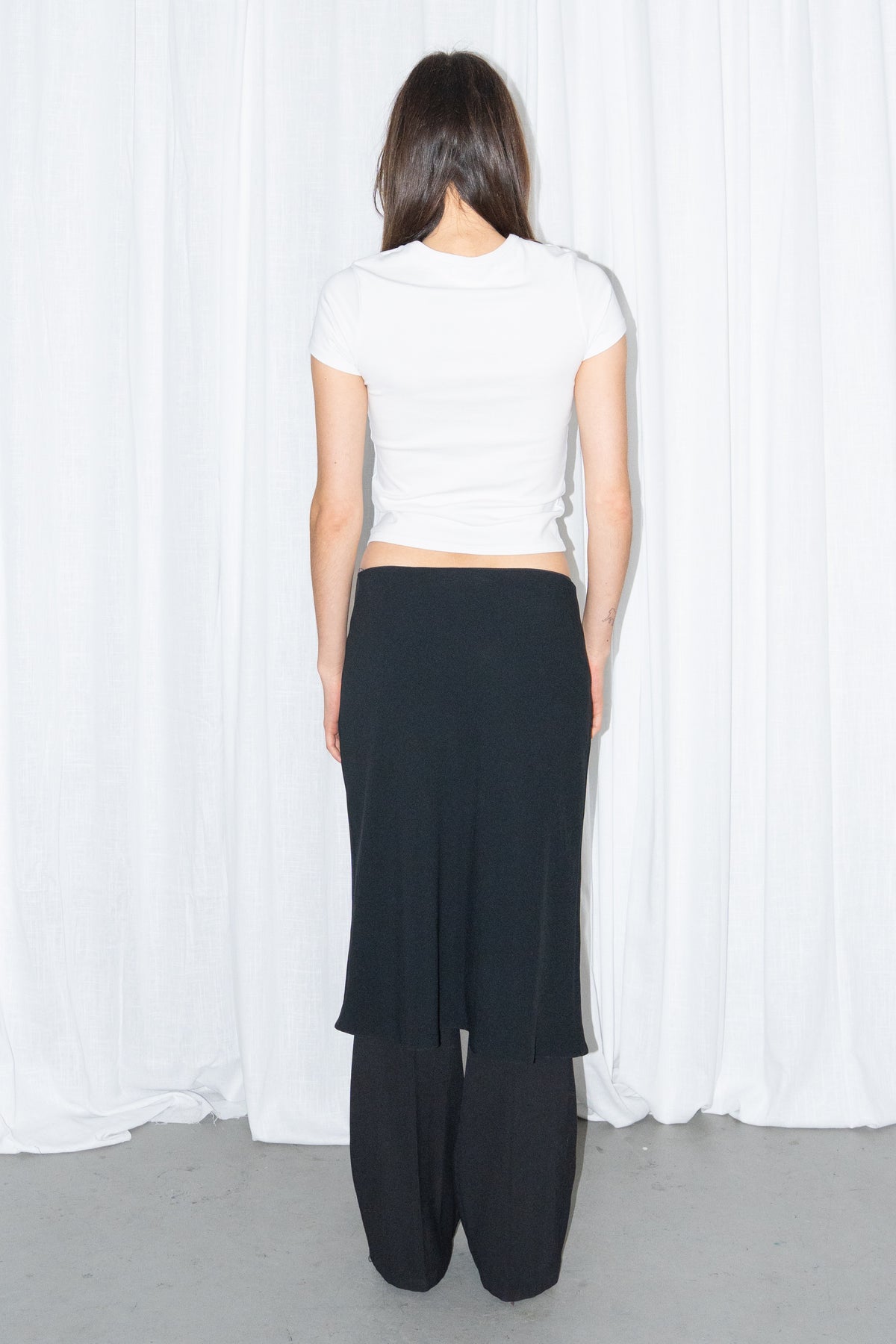 Back view of a white fitted T-shirt with a black flared midi skirt over black trousers