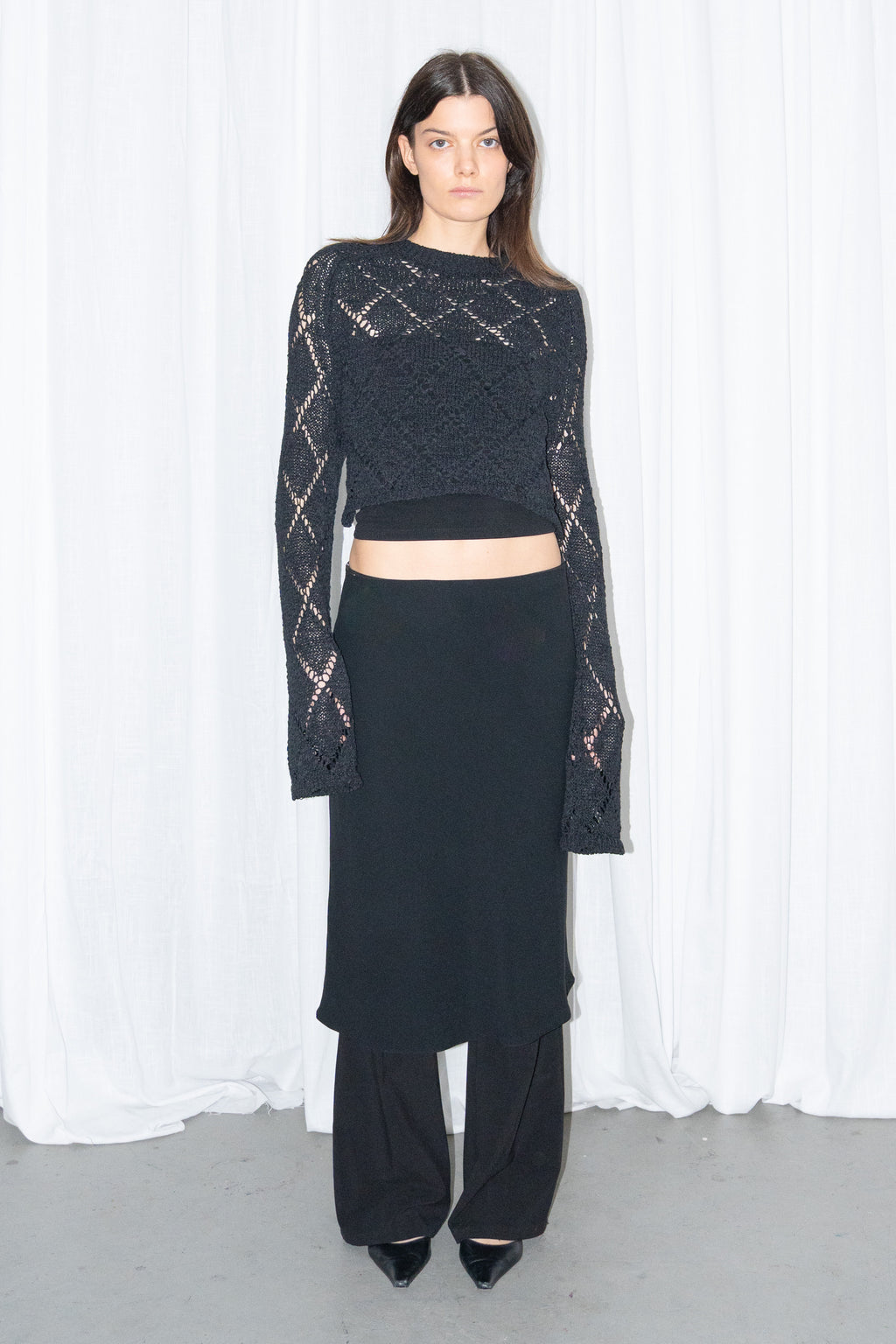Model wearing a black back cut-out crochet-style knit top with a black flared midi skirt over black trousers
