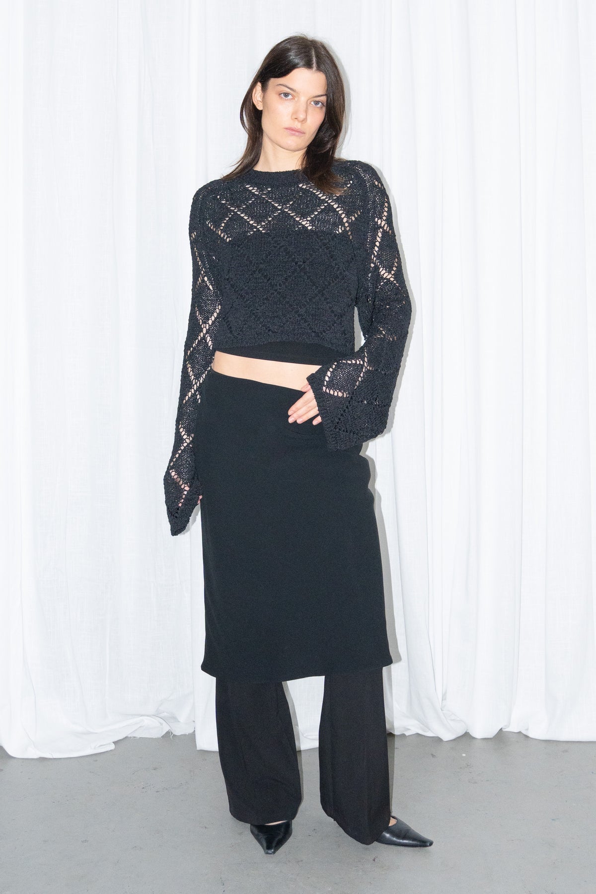 Model wearing a black back cut-out crochet-style knit top with a black flared midi skirt over black trousers