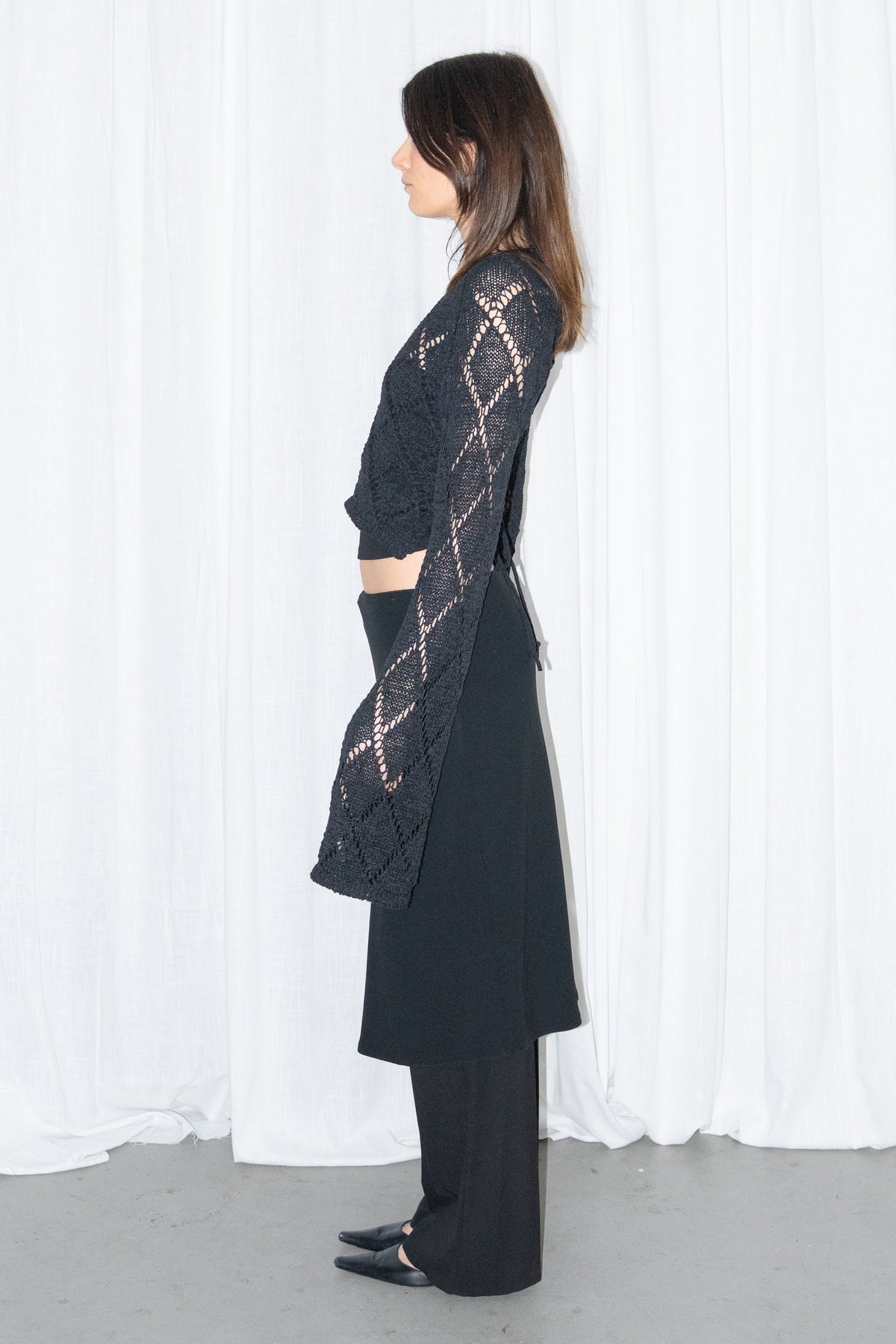 Model wearing a black back cut-out crochet-style knit top with a black flared midi skirt over black trousers