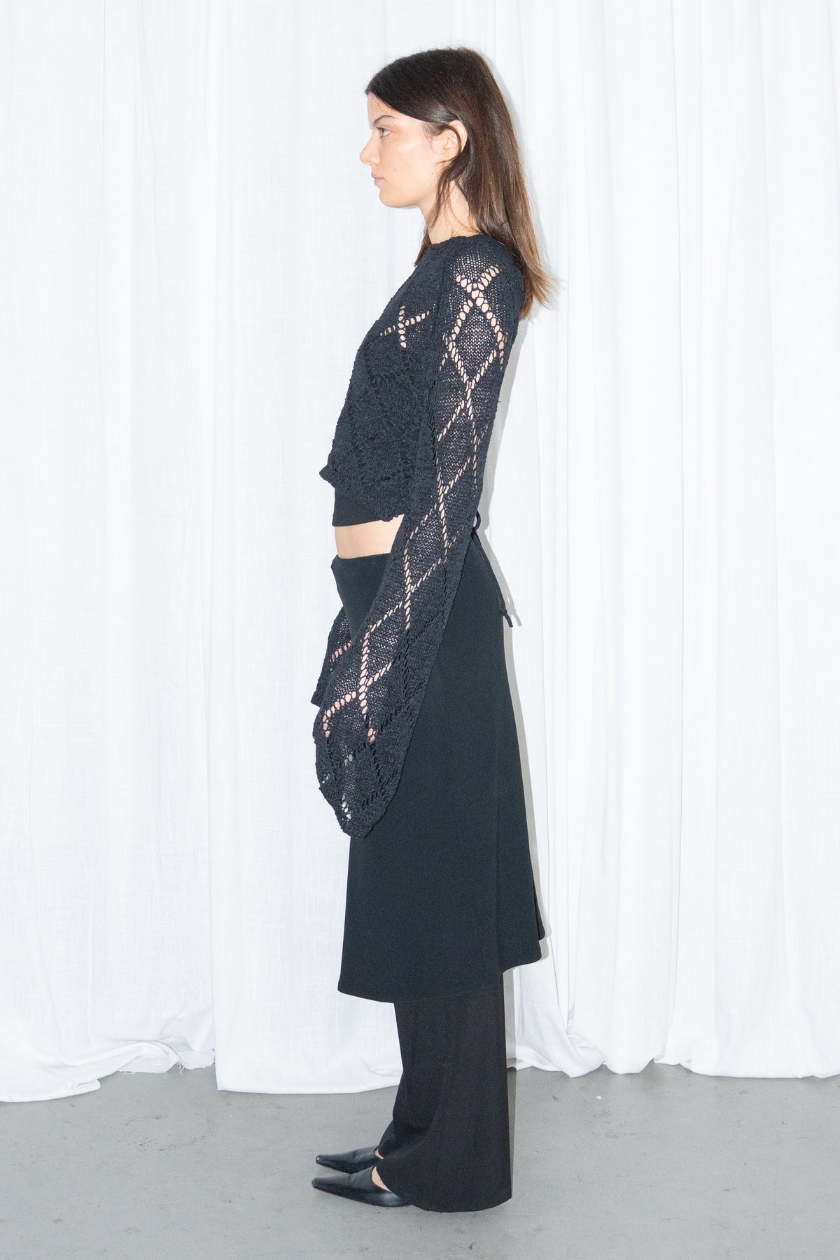 Model wearing a black back cut-out crochet-style knit top with a black flared midi skirt over black trousers