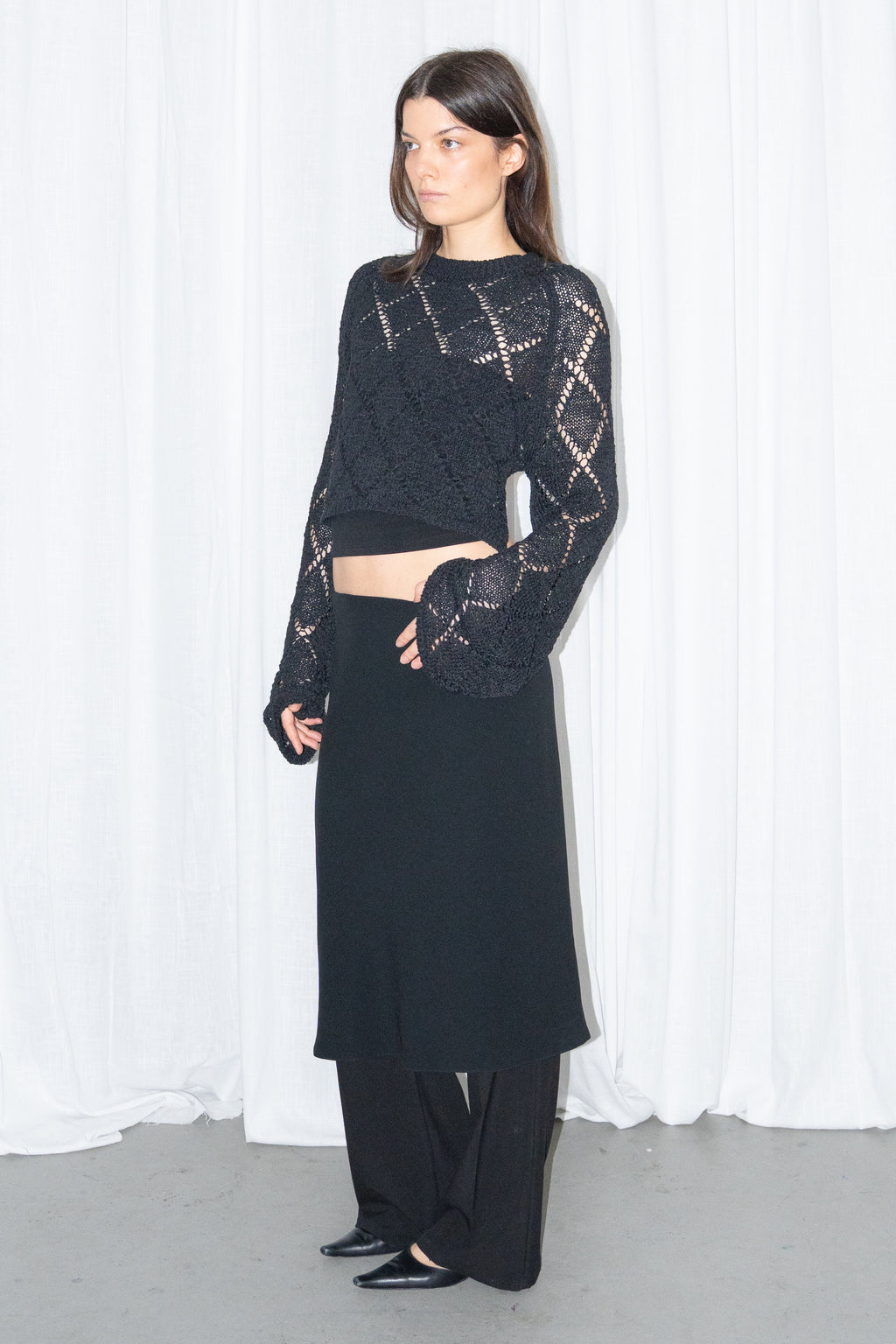 Model wearing a black back cut-out crochet-style knit top with a black flared midi skirt over black trousers