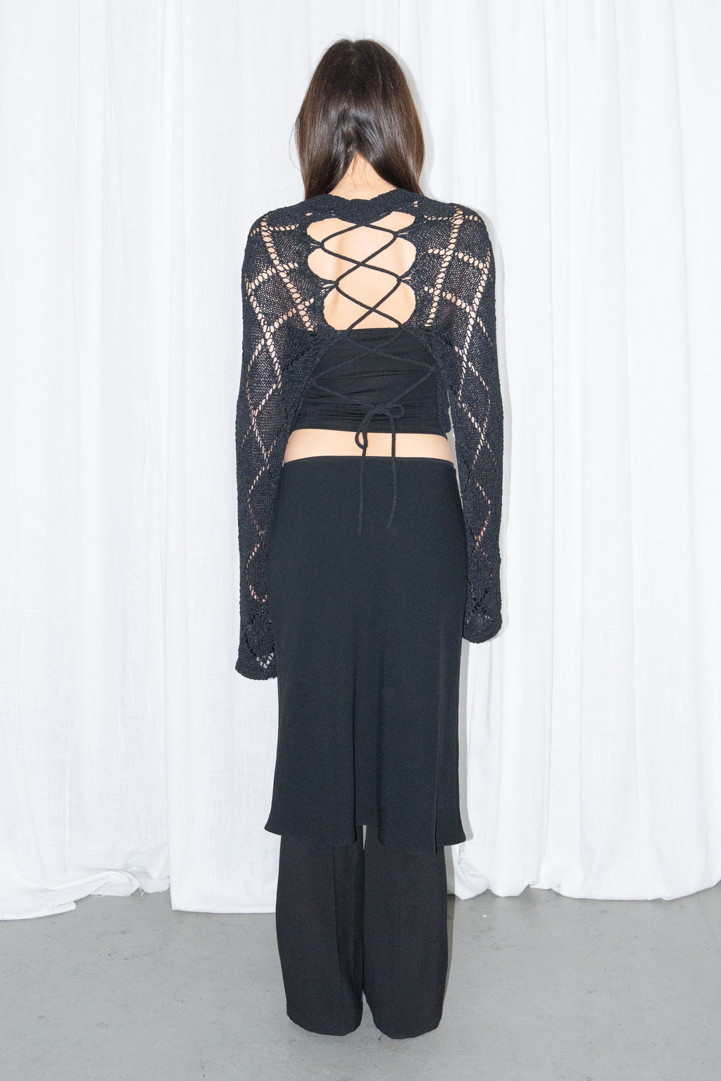 Back view of a black back cut-out crochet-style knit top with a black flared midi skirt over black trousers