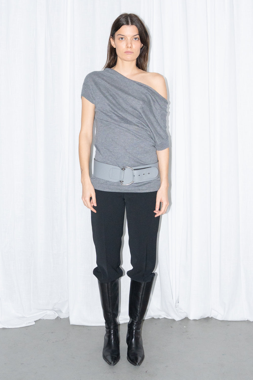 Model wearing a grey off-shoulder draped jersey knit top with a grey leather belt and black high-waist trousers