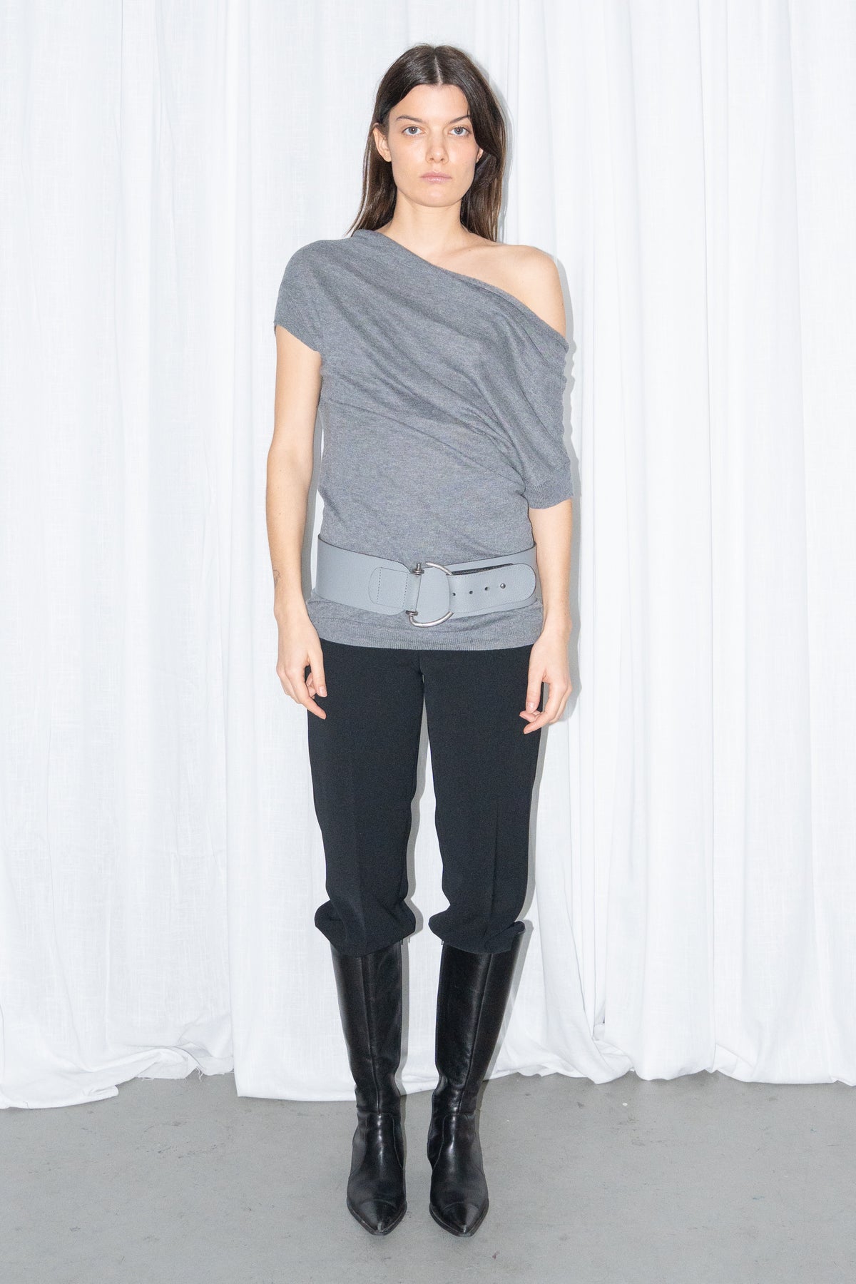 Model wearing a grey off-shoulder draped jersey knit top with a grey leather belt and black high-waist trousers