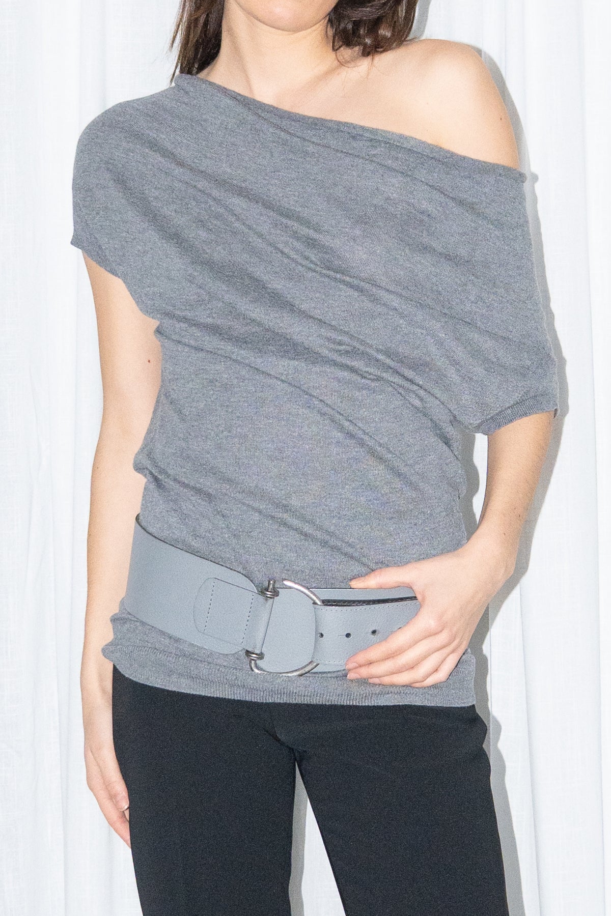 Close-up of a grey off-shoulder draped jersey knit top with a grey leather belt and black high-waist trousers