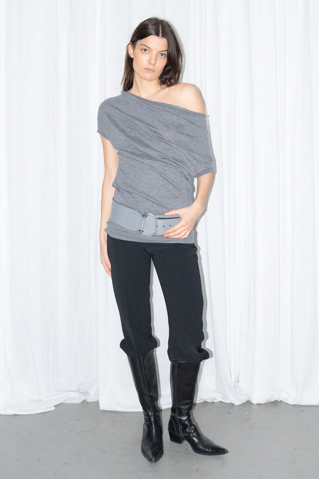 Model wearing a grey off-shoulder draped jersey knit top with a grey leather belt and black high-waist trousers