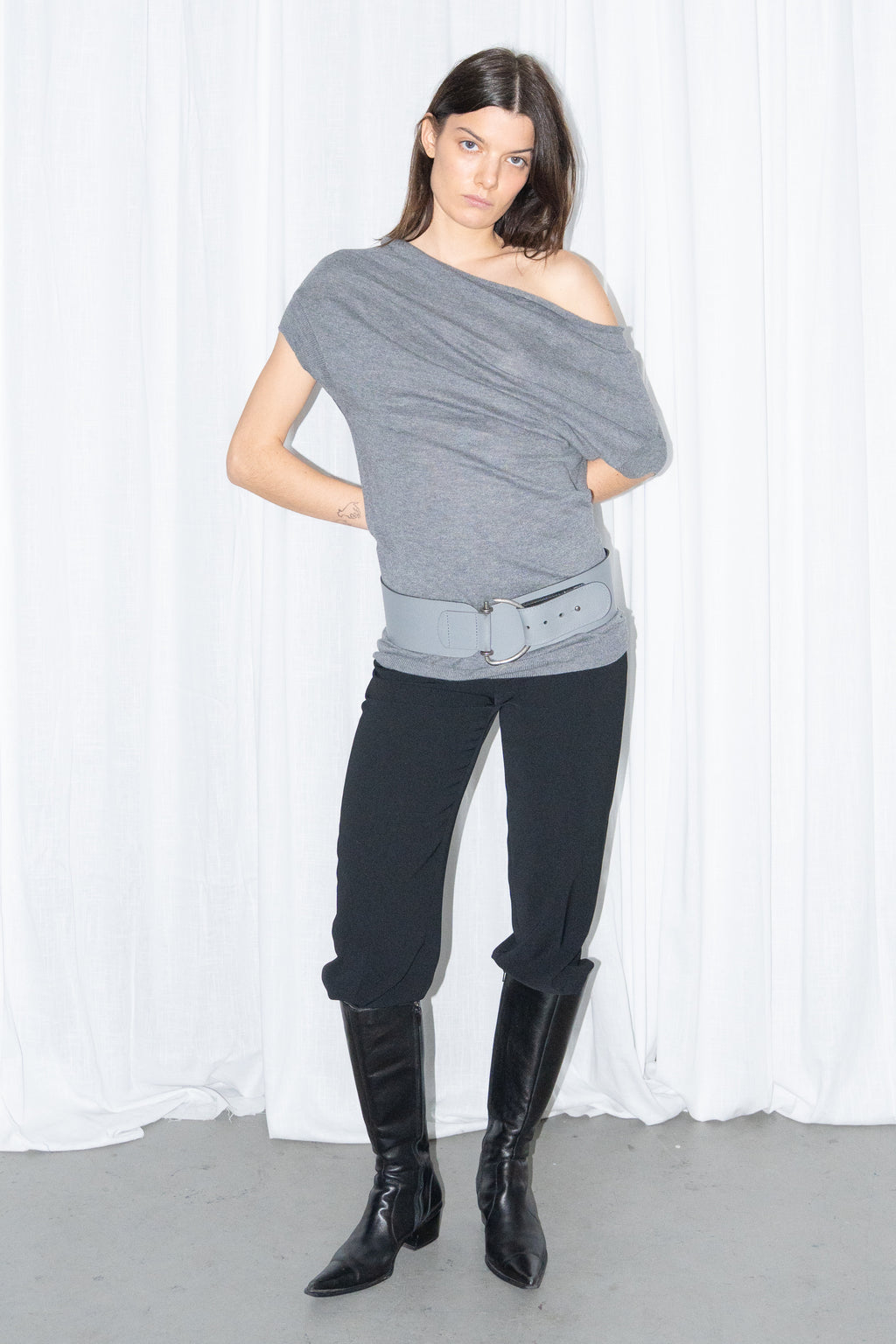 Model wearing a grey off-shoulder draped jersey knit top with a grey leather belt and black high-waist trousers