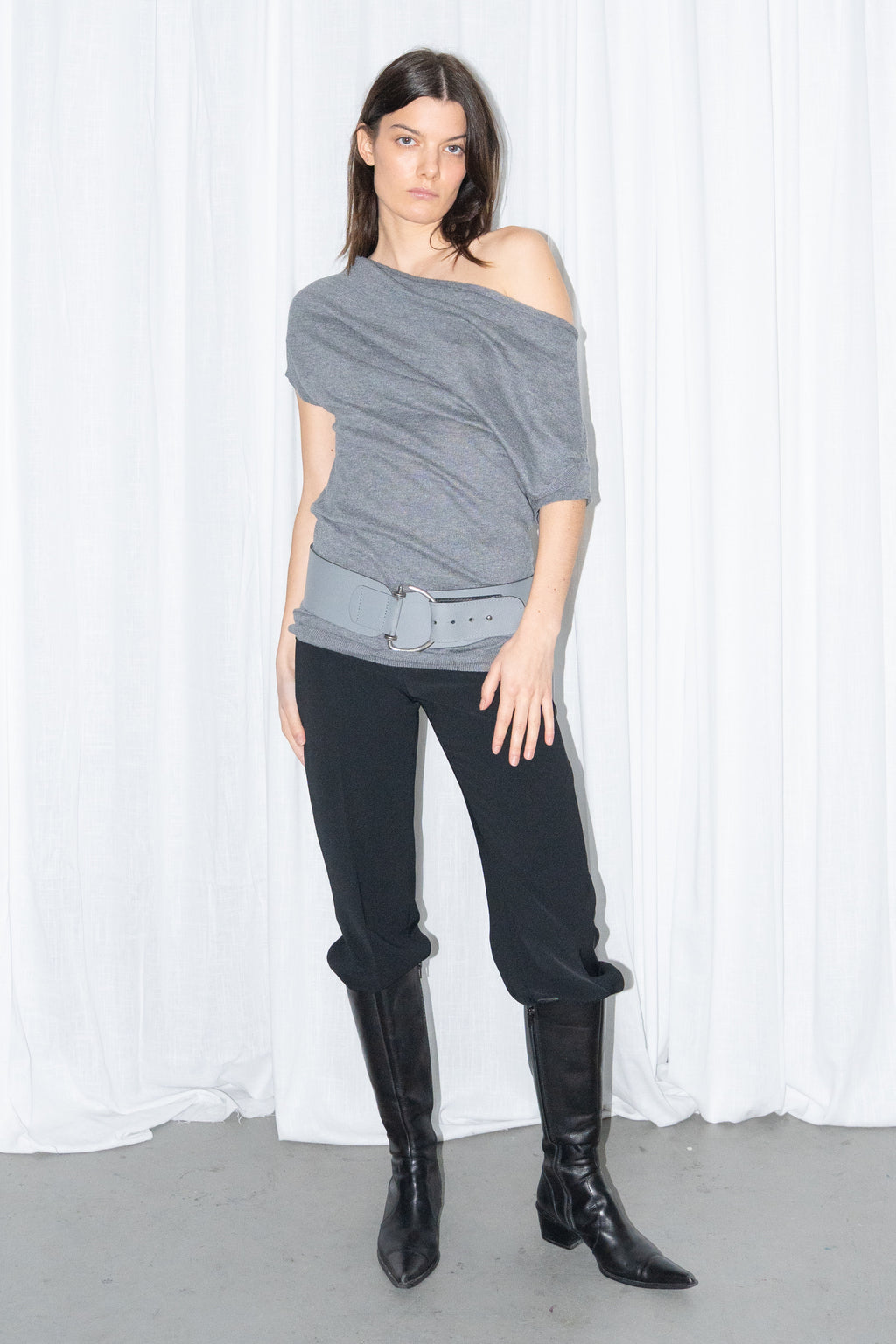 Model wearing a grey off-shoulder draped jersey knit top with a grey leather belt and black high-waist trousers