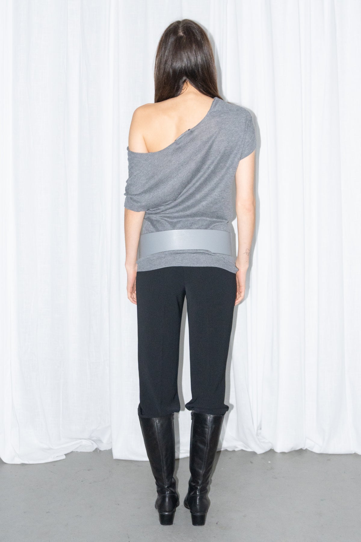 Back view of a grey off-shoulder draped jersey knit top with a grey leather belt and black high-waist trousers