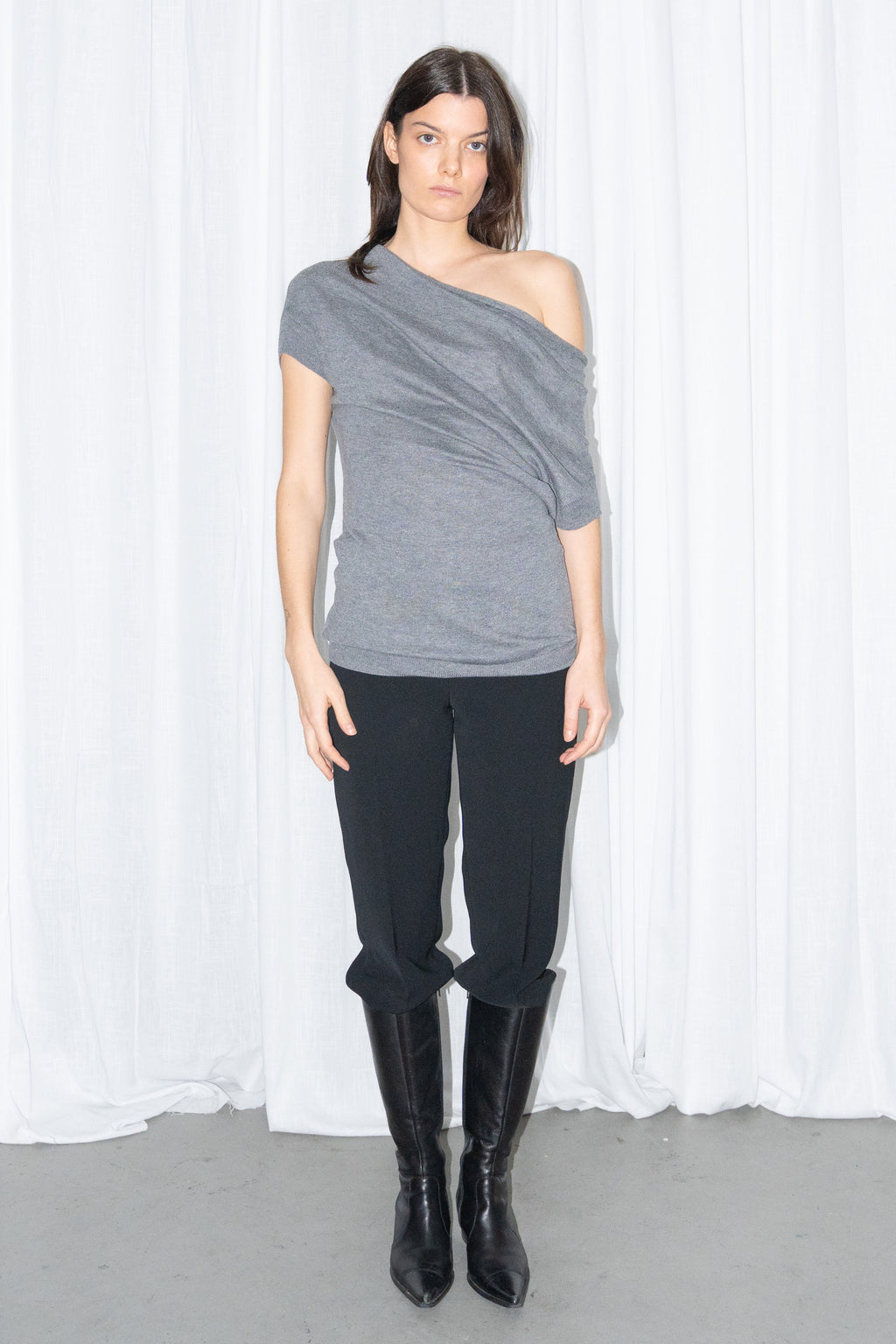 Model wearing a grey off-shoulder draped jersey knit top with a grey leather belt and black high-waist trousers