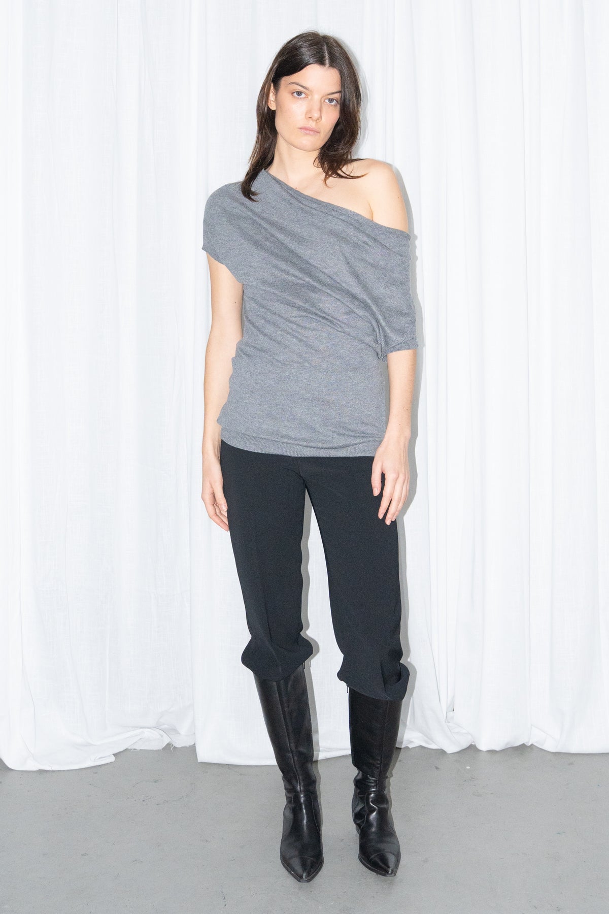 Model wearing a grey off-shoulder draped jersey knit top with a grey leather belt and black high-waist trousers