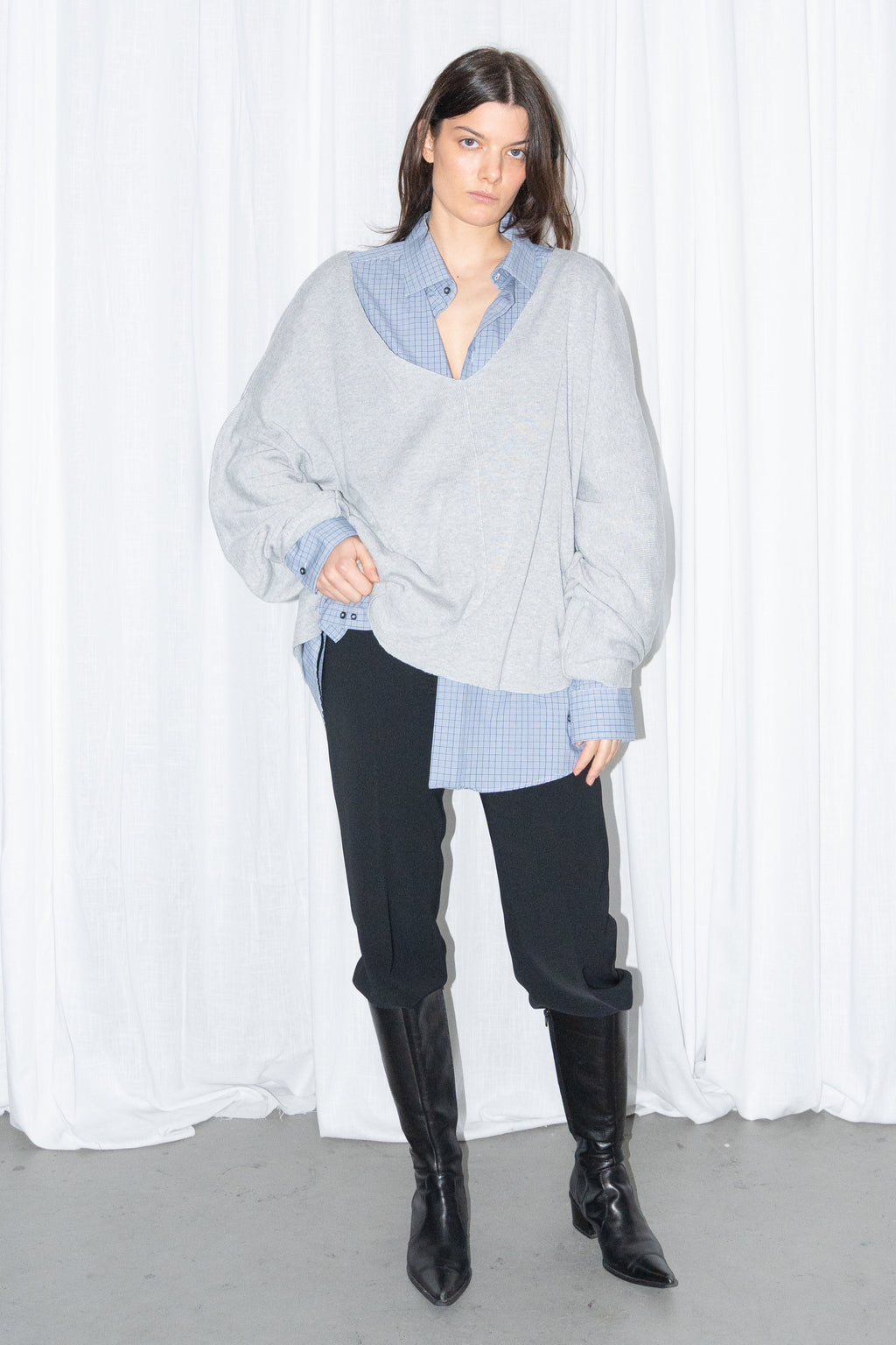 Model posing in a light grey oversized V-neck knit sweater over a light blue shirt with black jeans and knee boots