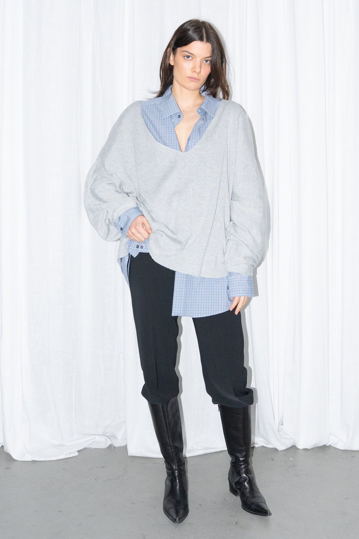 Model posing in a light grey oversized V-neck knit sweater over a light blue shirt with black jeans and knee boots