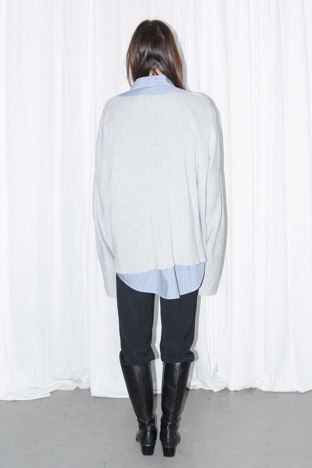 Back view of a light grey oversized V-neck knit sweater over a light blue shirt with black jeans and knee boots