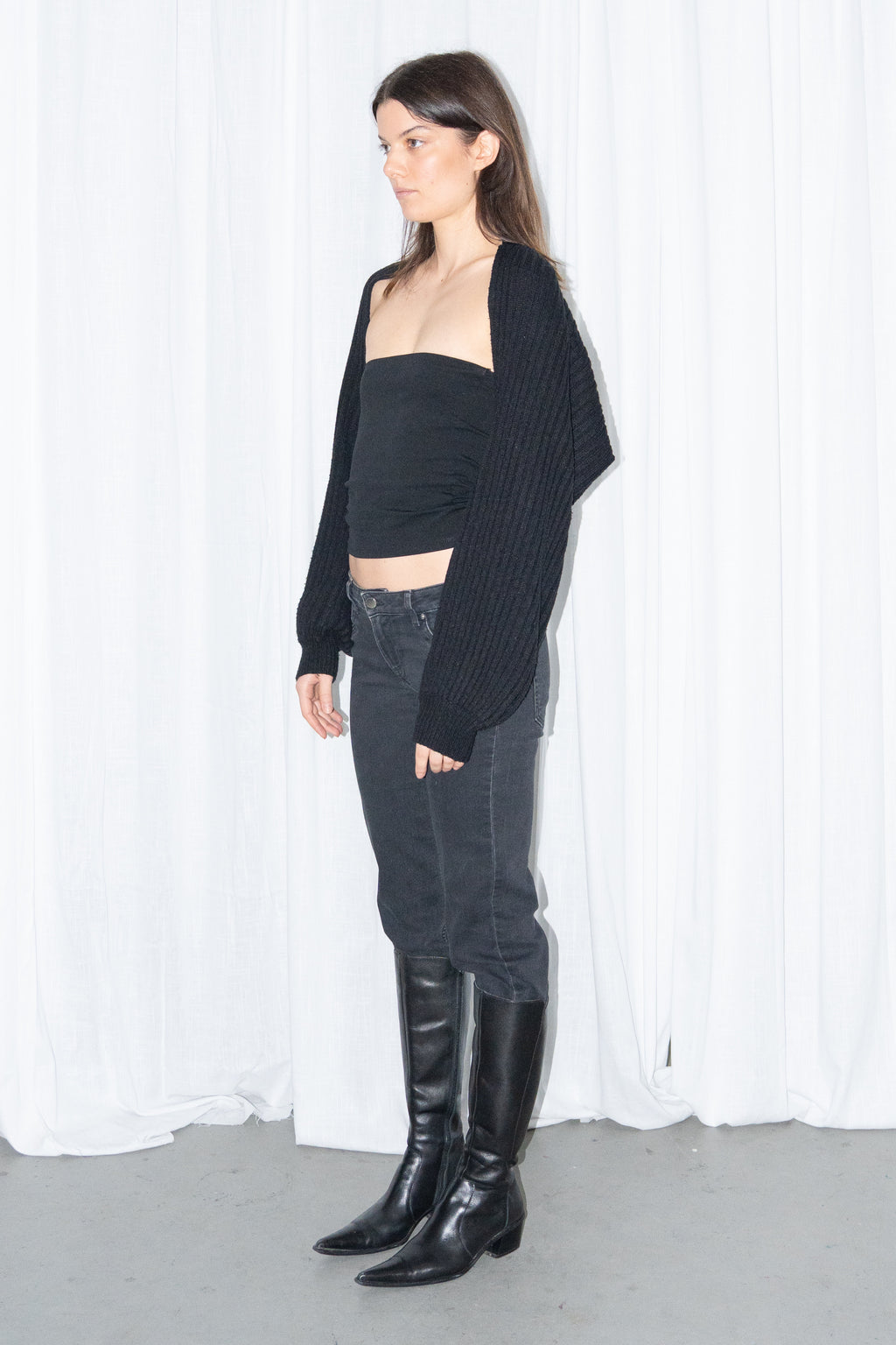 Side view of a black chunky knit cropped cardigan with a black bandeau top and black low-rise straight jeans